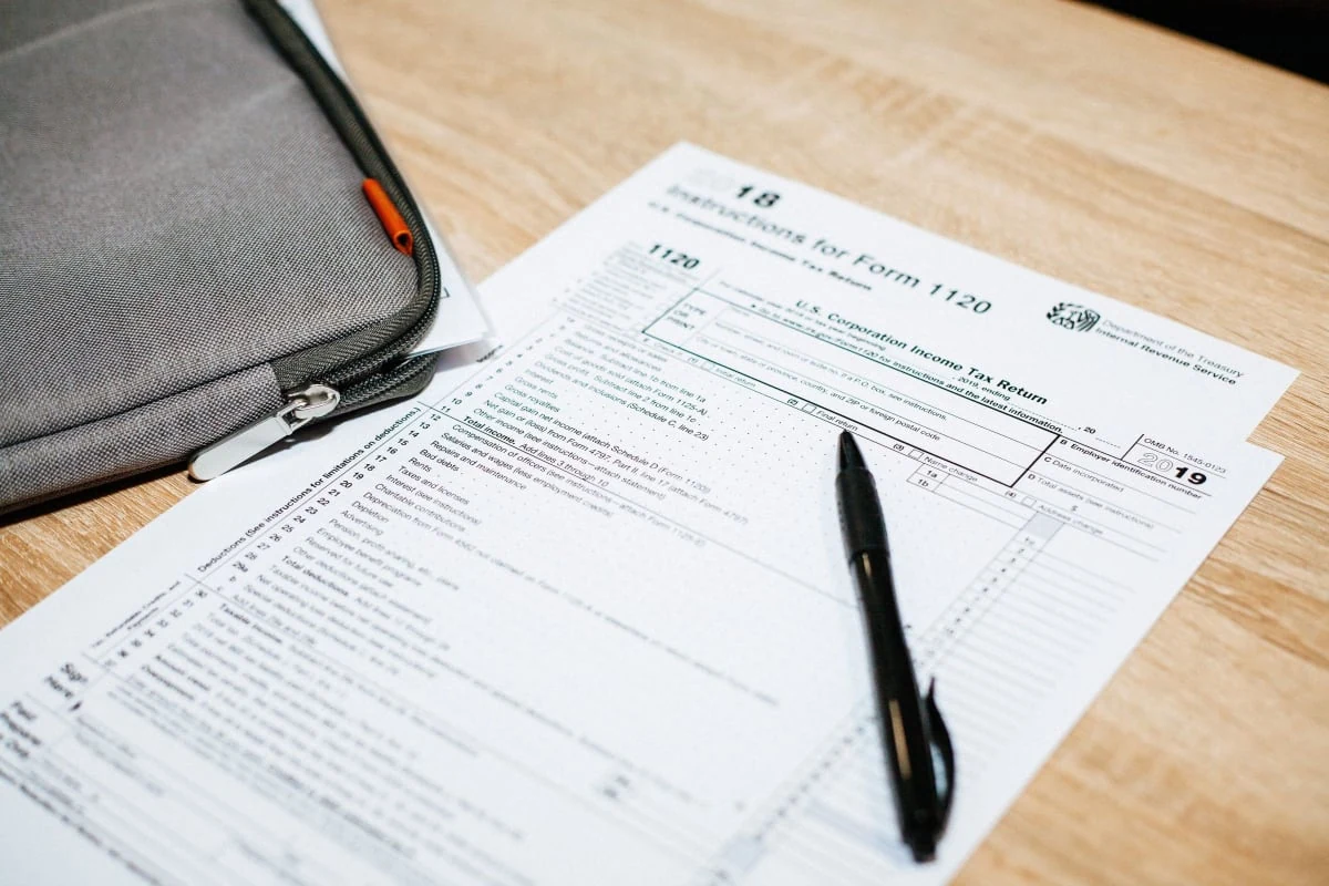 Irs form 1120 for a Tax Preparation Service in Nepean on a wooden desk with a black pen on top, beside a gray zippered bag.