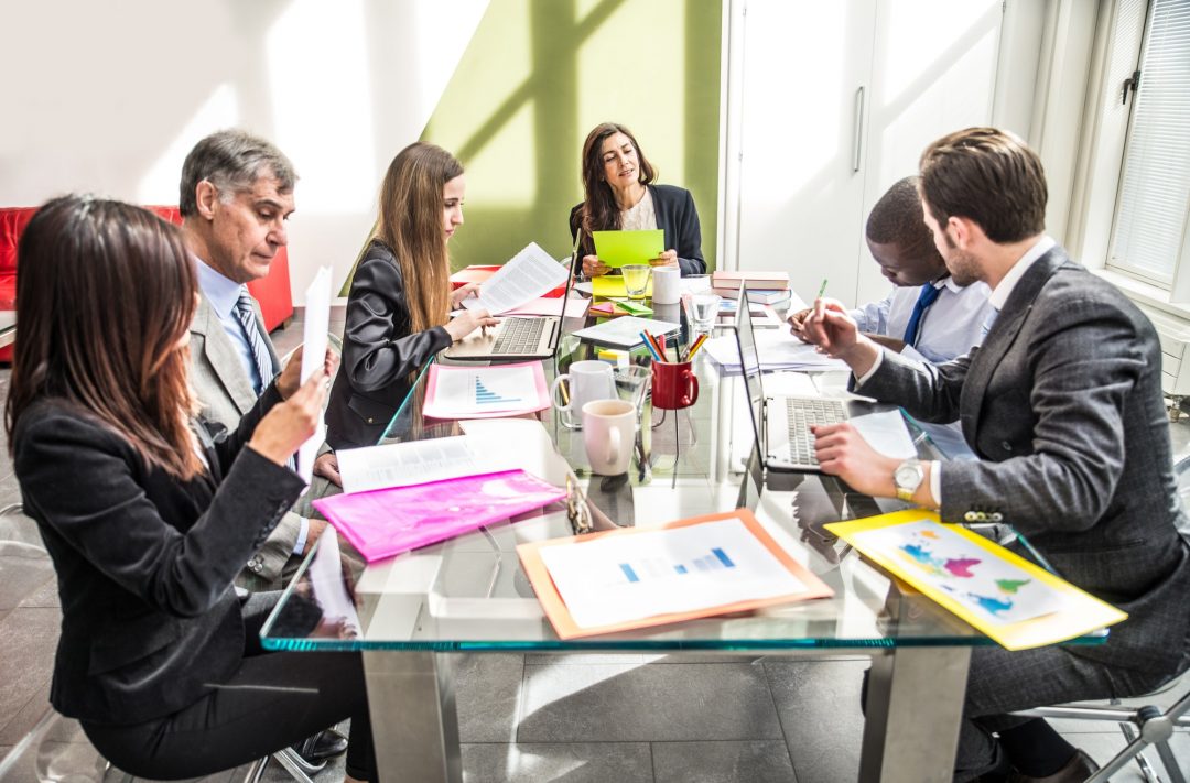 Six colleagues in formal attire are seated around a glass table in a brightly lit office, engaged in a meeting with documents and laptops, discussing bookkeeping strategies for their Ottawa clientele.