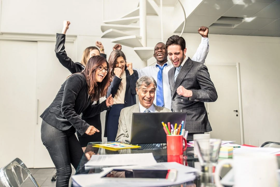 A group of six people in business attire cheer around a laptop in an office setting, showing excitement and happiness after finding top-notch bookkeeping services in Ottawa.
