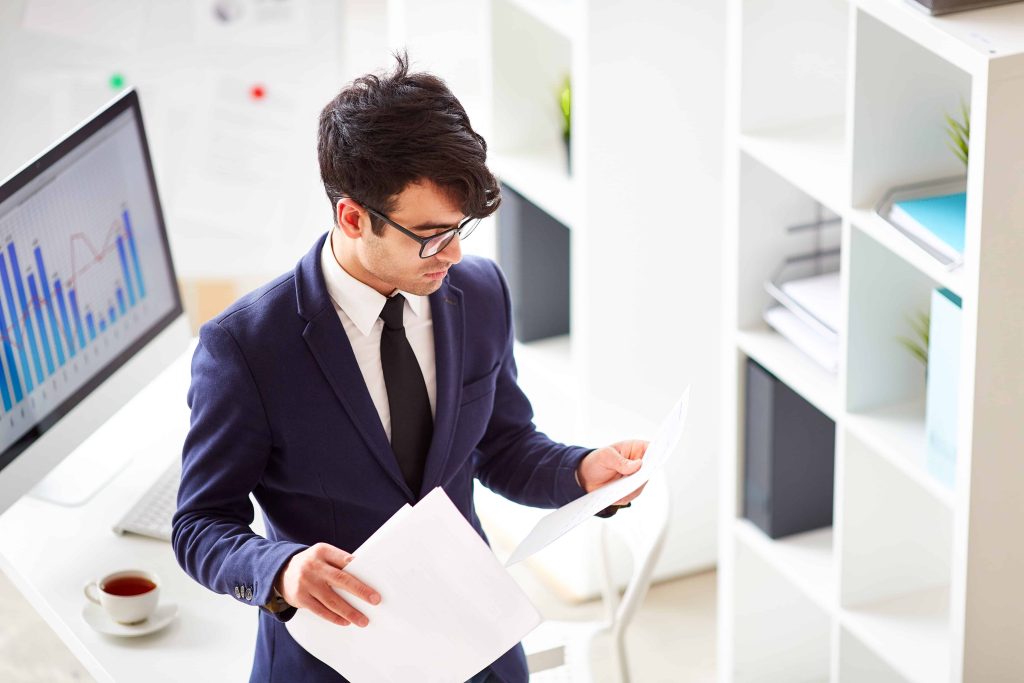 A tax accountant in Ottawa, dressed in a navy blue suit and black tie, stands in an office, meticulously reviewing documents. A computer displaying a bar graph rests on the desk beside him, with bookshelves lining the background.