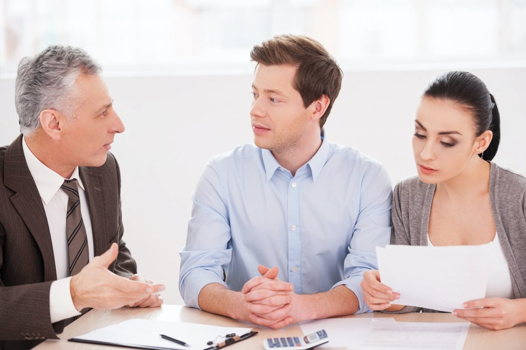 Three people are sitting at a table, having a discussion. The person on the left is gesturing while speaking, the person in the middle is listening intently, and the person on the right is holding and reading a document related to bookkeeping in Ottawa.