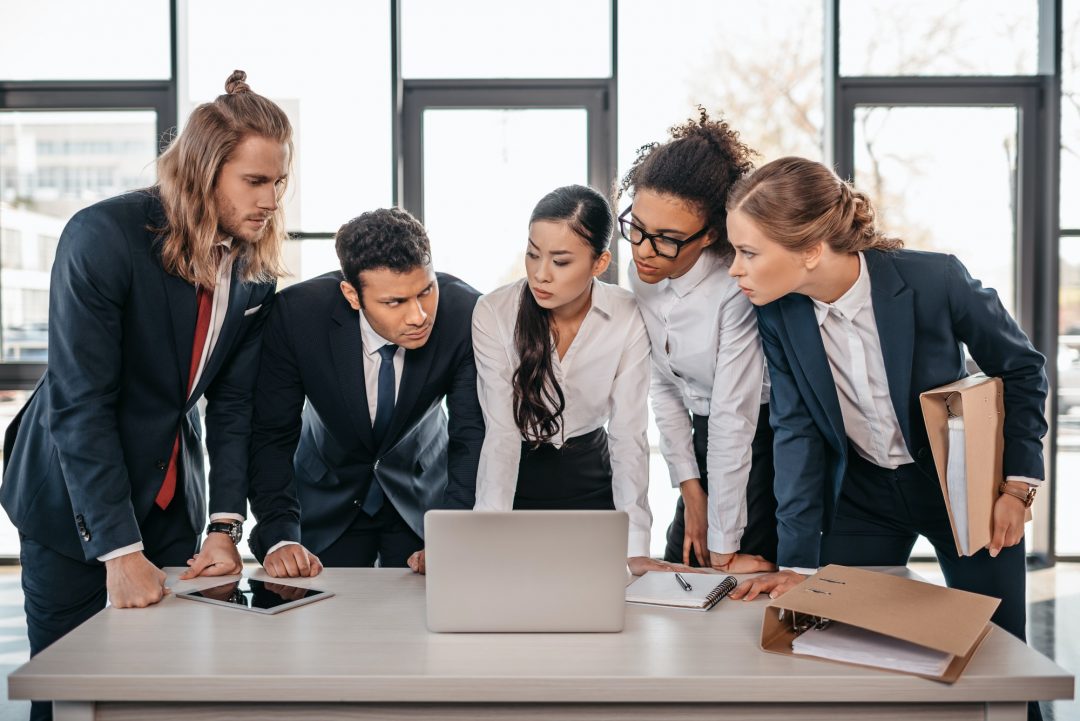 A diverse group of five business professionals, dressed in formal attire, closely examining a laptop screen in a modern office setting, discussing Income Tax Services in Ottawa.