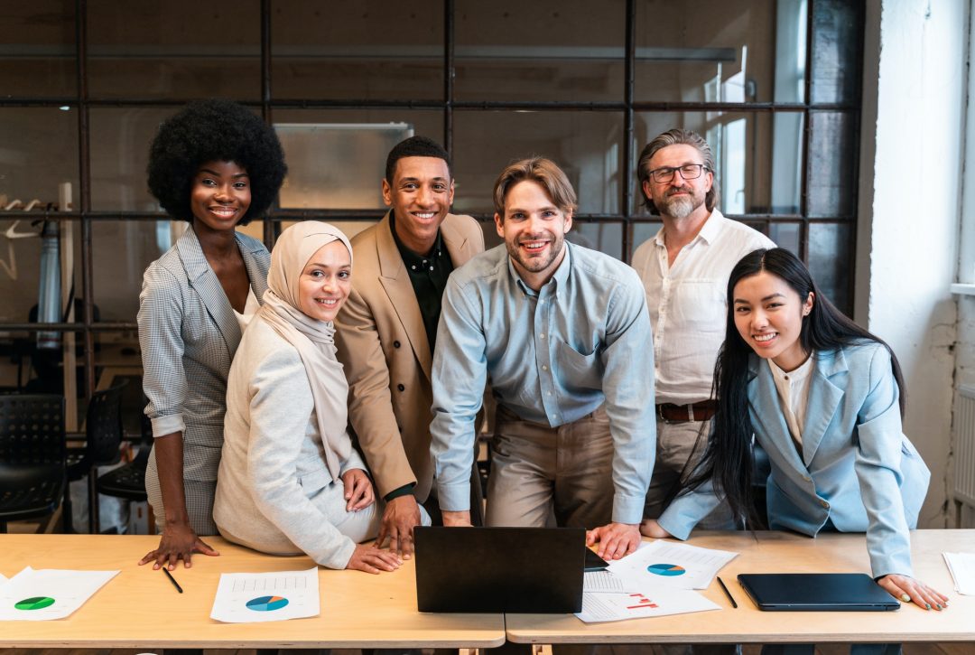 Six professionally dressed people, including a chartered accountant in Ottawa, stand and smile around a table with documents, charts, and a laptop in a modern office setting.