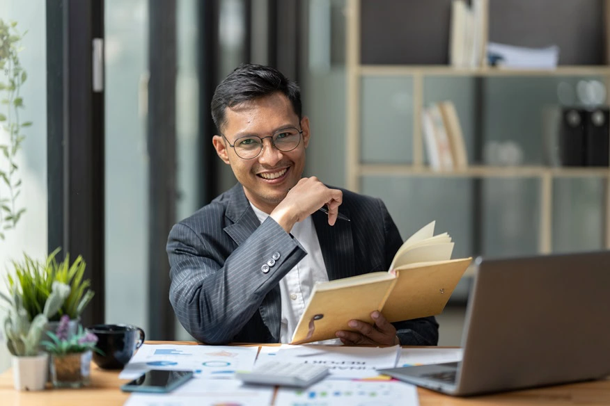 A Chartered Accountant in Ottawa sits at a desk with an open book, smiling. He wears a suit and glasses. The desk features a laptop, documents, and a potted plant. Bookshelves and large windows provide an elegant backdrop.