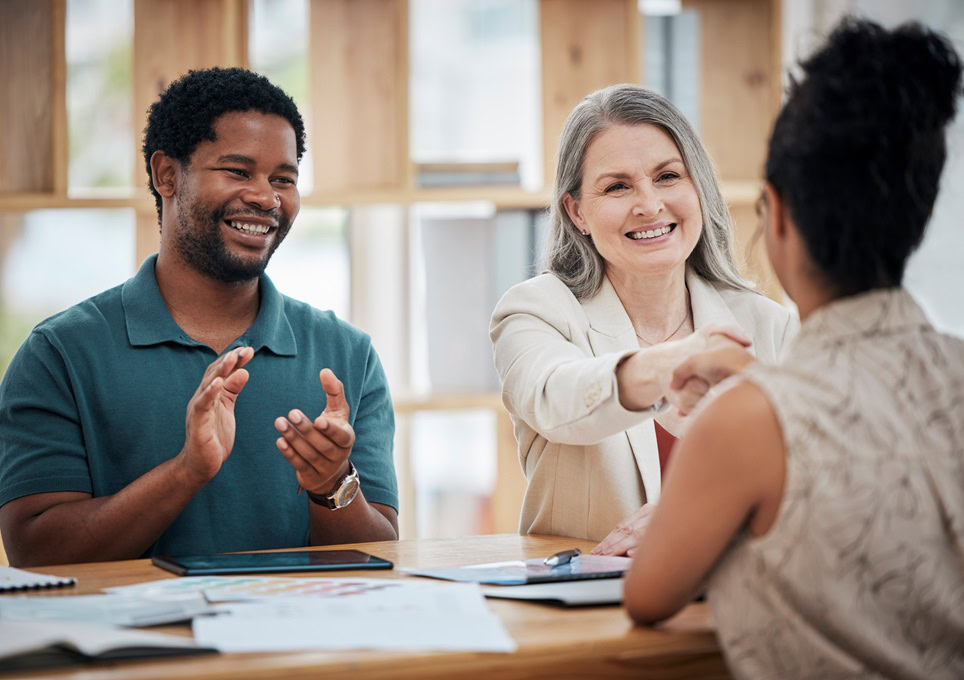 Three people in a meeting: a woman with gray hair shakes hands with a Tax Accountant in Ottawa facing away, while a man with a beard and short curly hair claps. Documents and tablets are on the table.
