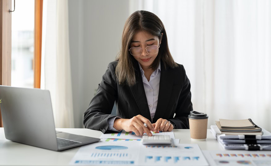 A Chartered Accountant in Ottawa sits at a desk using a calculator, surrounded by charts, documents, a laptop, and a coffee cup.