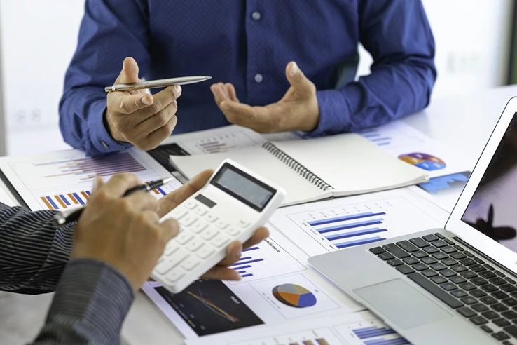 Two people, including a Chartered Accountant in Ottawa, are discussing financial charts and reports. One holds a pen while the other uses a calculator. A laptop, graphs, and a notebook are also spread across the table.