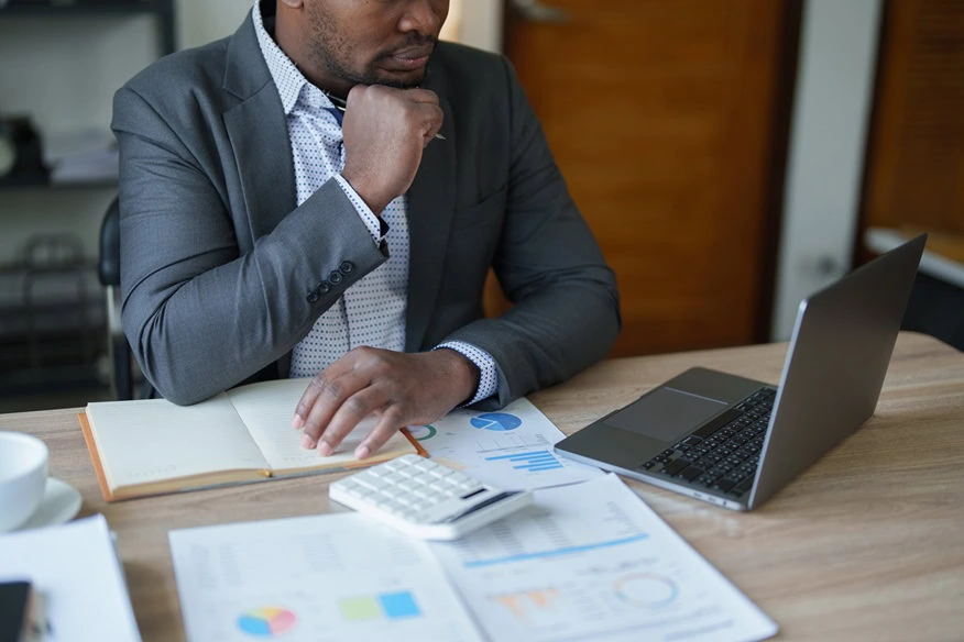 A Chartered Accountant in Ottawa sits at a desk with a laptop, open notebook, calculator, and documents displaying charts and graphs. They rest their chin on their hand, appearing deep in thought.