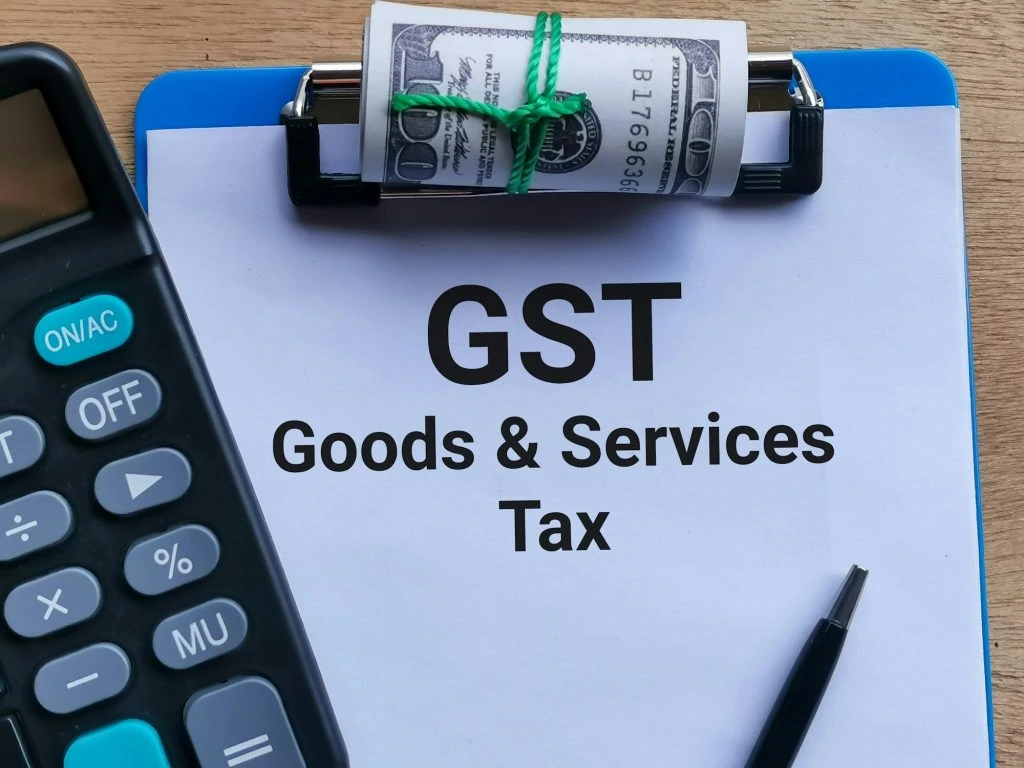 A clipboard with a paper titled "GST Goods & Services Tax," a rolled-up dollar bill secured with a green elastic band, a calculator, and a pen is essential for navigating Tax Services in Ottawa