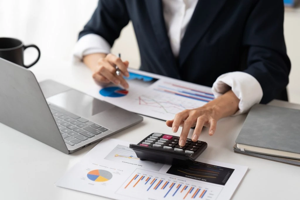 A person in a suit uses a calculator at a desk with a laptop, financial charts, documents, and a coffee mug, diligently preparing for Tax Services in Ottawa