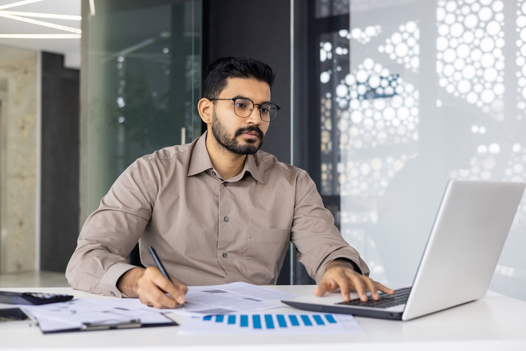 A Chartered Accountant in Ottawa, wearing glasses, sits at a desk working on a laptop, holding a pen and examining documents. Papers, a phone, and graphs are spread out on the desk. The background showcases modern office decor.