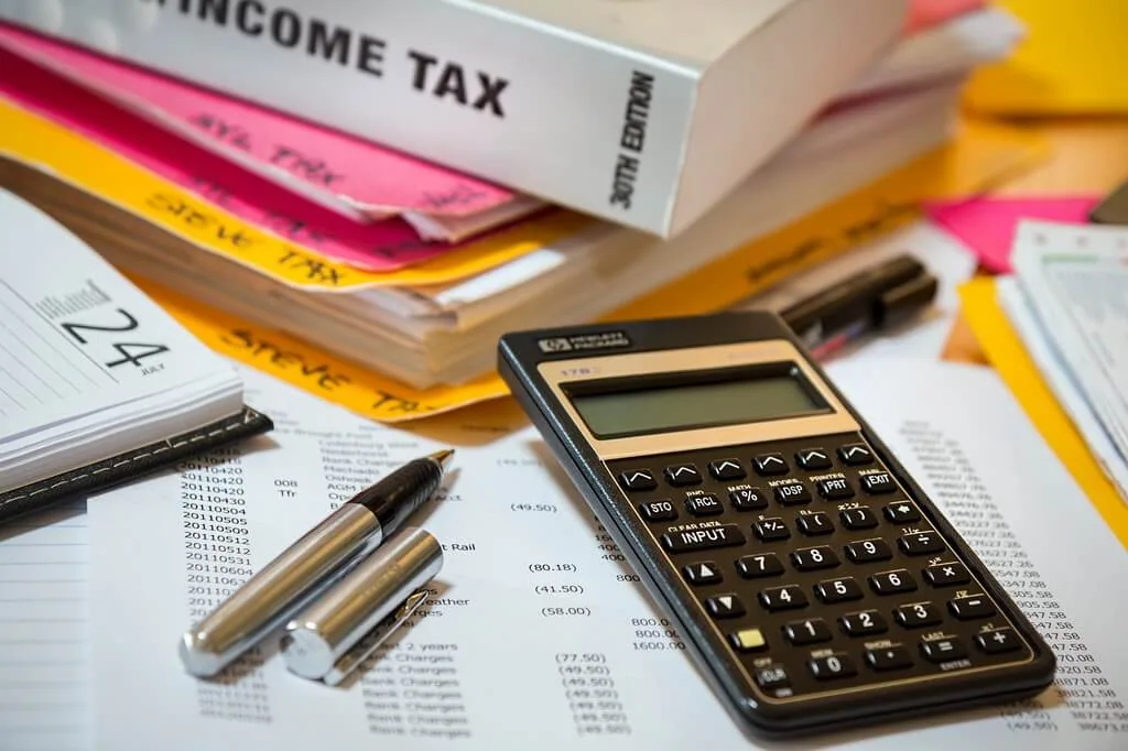 A cluttered desk with a calculator, pens, a tax form, stacks of folders, and an income tax reference book begs the question: How much does it cost to get your taxes done in Canada?