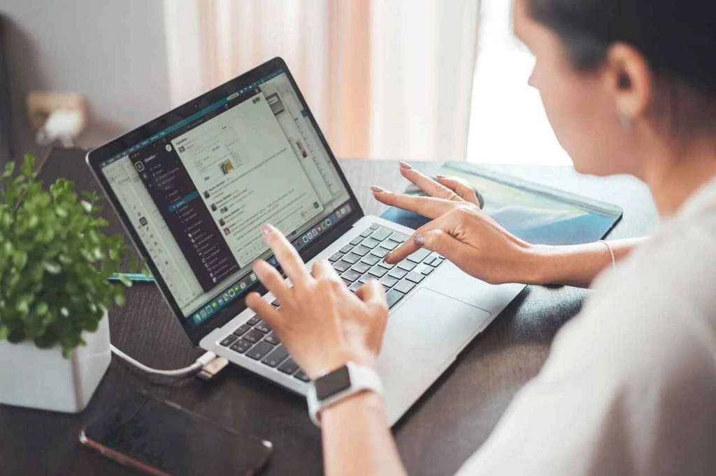 A person working on a laptop, utilizing various productivity tools, manages their tasks efficiently. Beside the laptop is a smartphone and a plant, creating an organized workspace perfect for running a bookkeeping service.