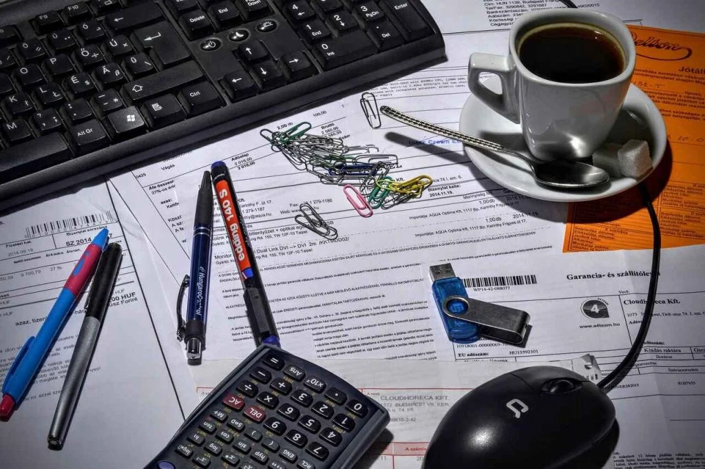 A cluttered desk with a keyboard, documents, coffee, calculator, pens, paper clips, USB drive, and a computer mouse - the quintessential setup for an efficient bookkeeping service.