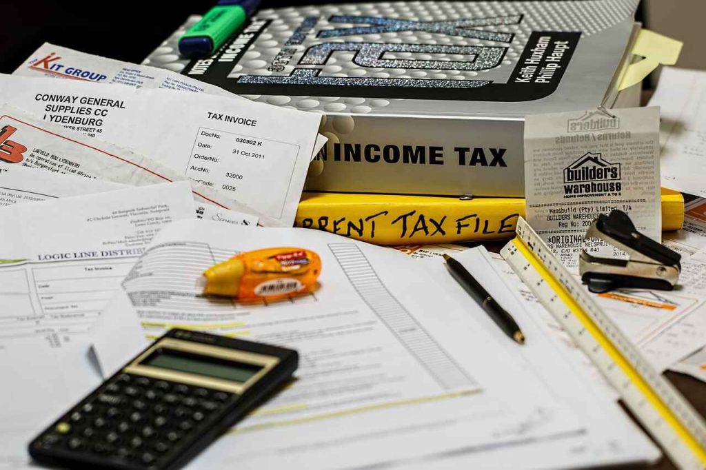 A cluttered desk with a calculator, tax documents, invoices, a "Current Tax File" folder, a book titled "Income Tax," pens, a pencil, a highlighter, and other office supplies suggests the busy life of an accountant preparing for tax return season.
