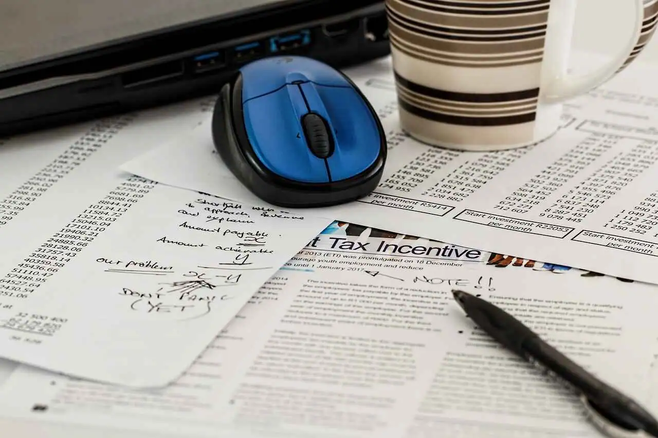 A blue computer mouse sits atop printed documents, beside a mug and a pen. The documents include handwritten notes and a tax form, presumably ready for the tax consultant's review.